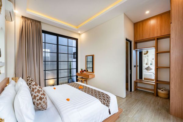 Cozy modern Balinese bedroom with king bed, wooden vanity, large grid windows, and warm lighting.